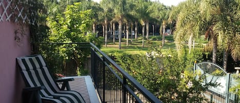 Ein Balkon mit einem Streifenstuhl, Blick auf einen Garten mit Palmen und ein Metallgitter.