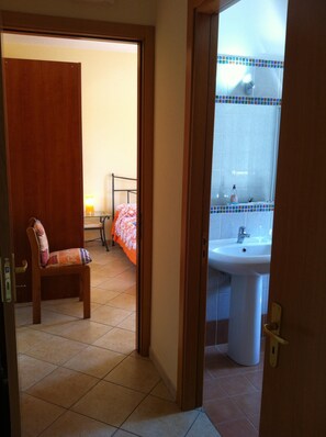 Una habitación de hotel con cama, una silla, baño con lavamanos y espejo, y piso de baldosas.
