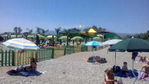 A beach with people sunbathing under colorful umbrellas, a green fence, and a mountain in the background.