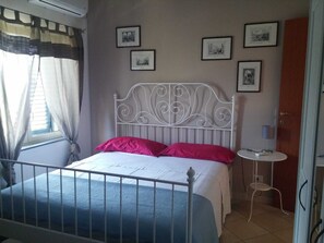 Un dormitorio con estructura de cama metálica, cabecera blanca, una mesita de noche y una silla. Cuatro cuadros enmarcados adornan la pared.