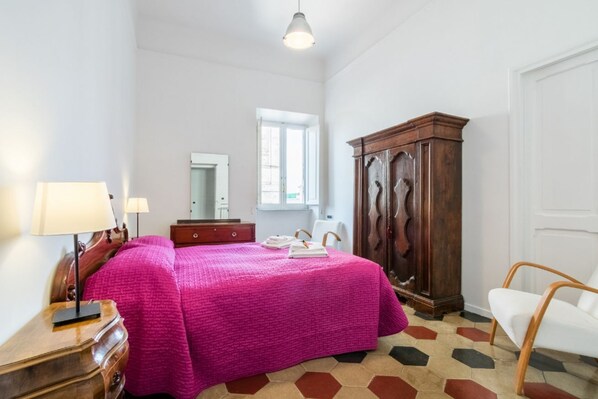 Quarto com uma cama, guarda-roupa de madeira, escrivaninha com abajur, cadeira e janela.