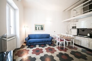Un ambiente compatto con un divano blu, un tavolo da pranzo con sedie, angolo cottura con pensili e pavimento a motivi.