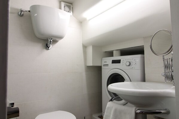A compact bathroom with a toilet, a sink, a washing machine, and a wall-mounted toilet paper dispenser.