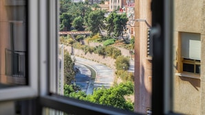 Vista pela janela de um edifício, mostrando um rio, vegetação e outros edifícios ao fundo.