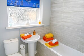 Et badeværelse med en hvid vask, toilet og badekar. Et vindue over vasken giver udsigt til sejlbåde.