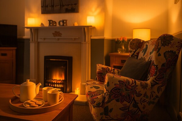 Un salón acogedor con chimenea, un sillón floral, una mesa con tetera, tazas y galletas, y una estantería de pared con adornos.