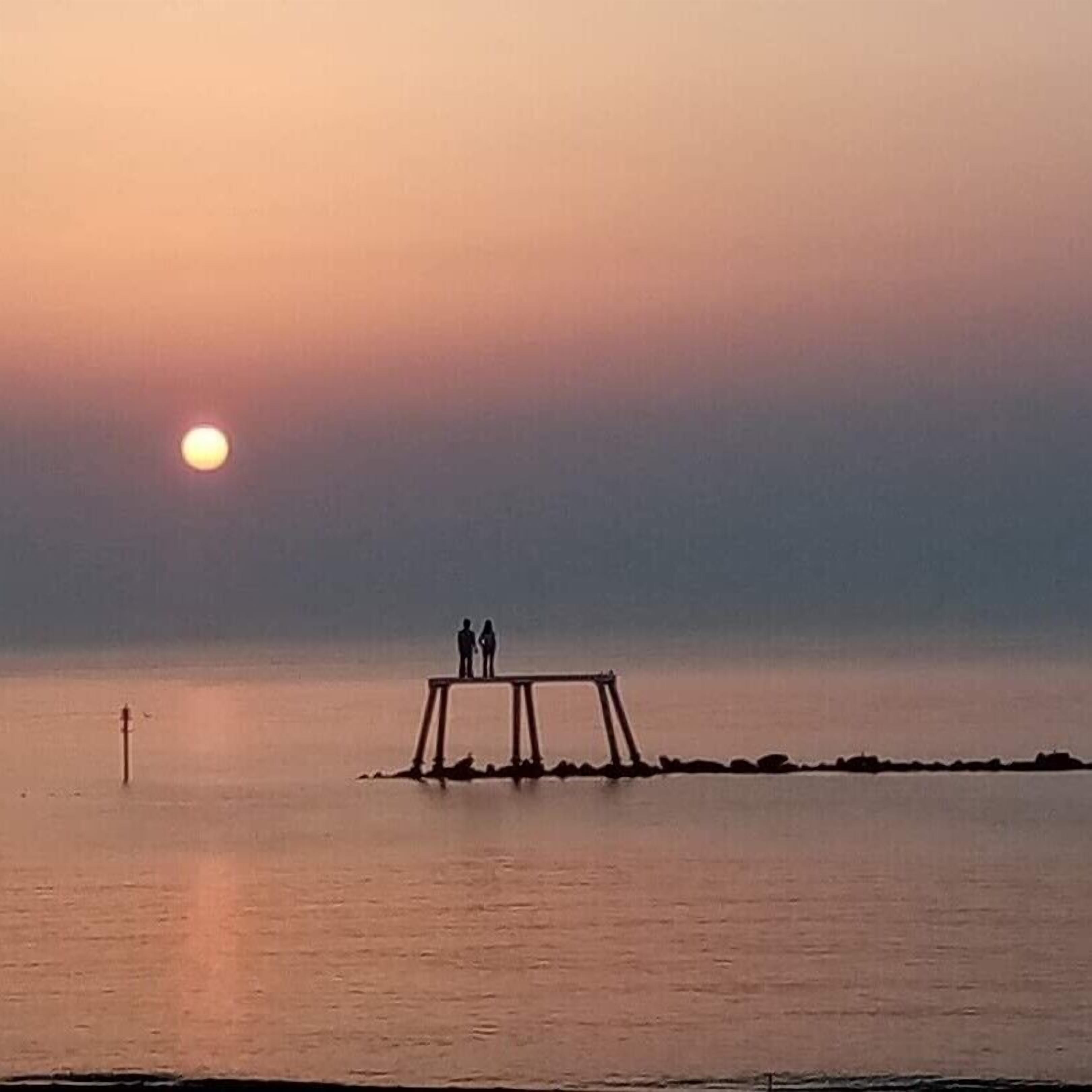 A sunset over a calm body of water with a wooden pier and two people standing on it.