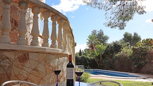 A table with a bottle of wine and two glasses, set against a stone balustrade overlooking a swimming pool and lush greenery.