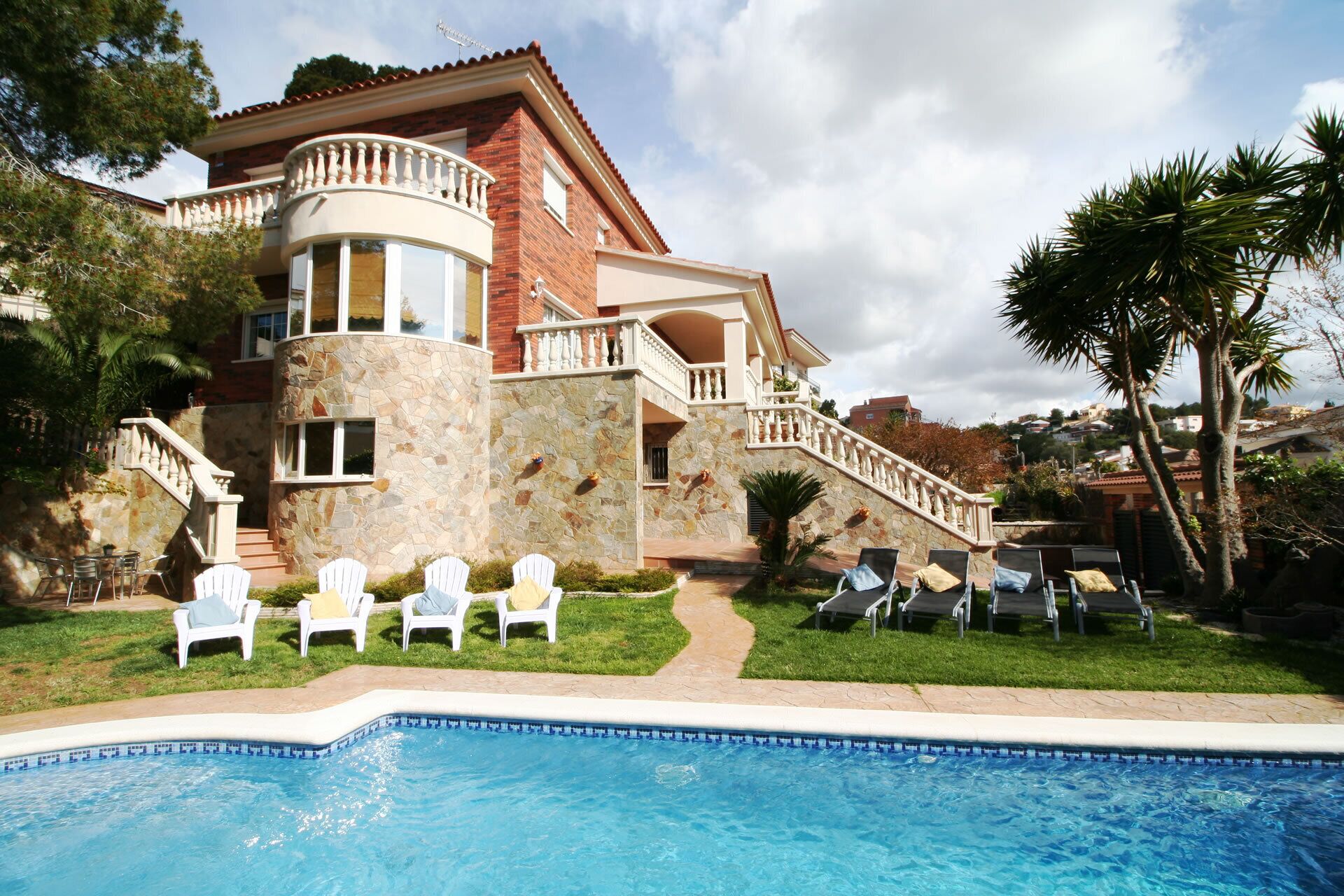 A two-story house with a stone facade, a pool, and white outdoor chairs.