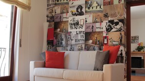 A living room with a white sofa, red and grey cushions, and a wall covered with various posters and artwork.