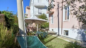 Una hamaca en un patio trasero con una mesa y sillas, rodeado de follaje y un edificio.