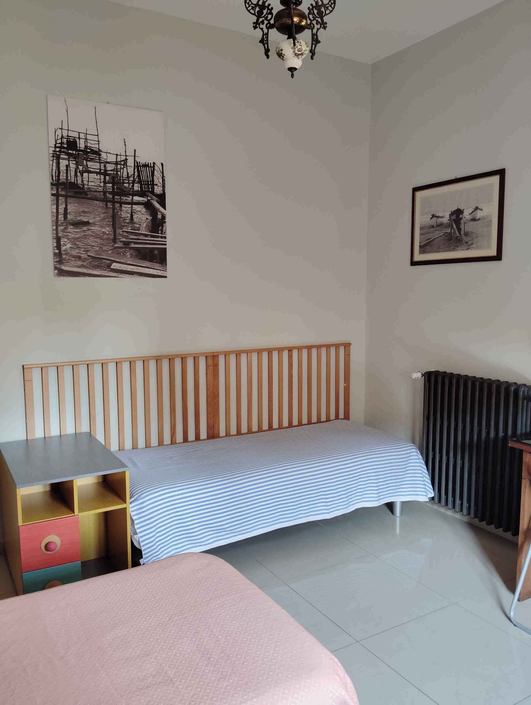 Een kamer met een houten bankje, een bed met een gestreept dekbedovertrek, een radiator, een klein tafeltje met lade en twee ingelijste foto's aan de muur.