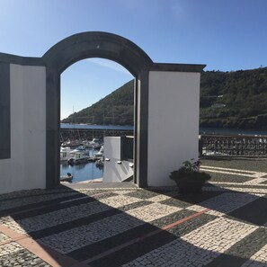 Uitzicht door een boogvormige deuropening op een jachthaven met boten, met bergen op de achtergrond en een verhard gebied met een plant in pot.