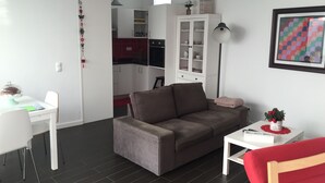 A modern living room with a grey sofa, a red armchair, a white desk, and a kitchen area in the background. - Traditional Angra House (Angra do Heroísmo)