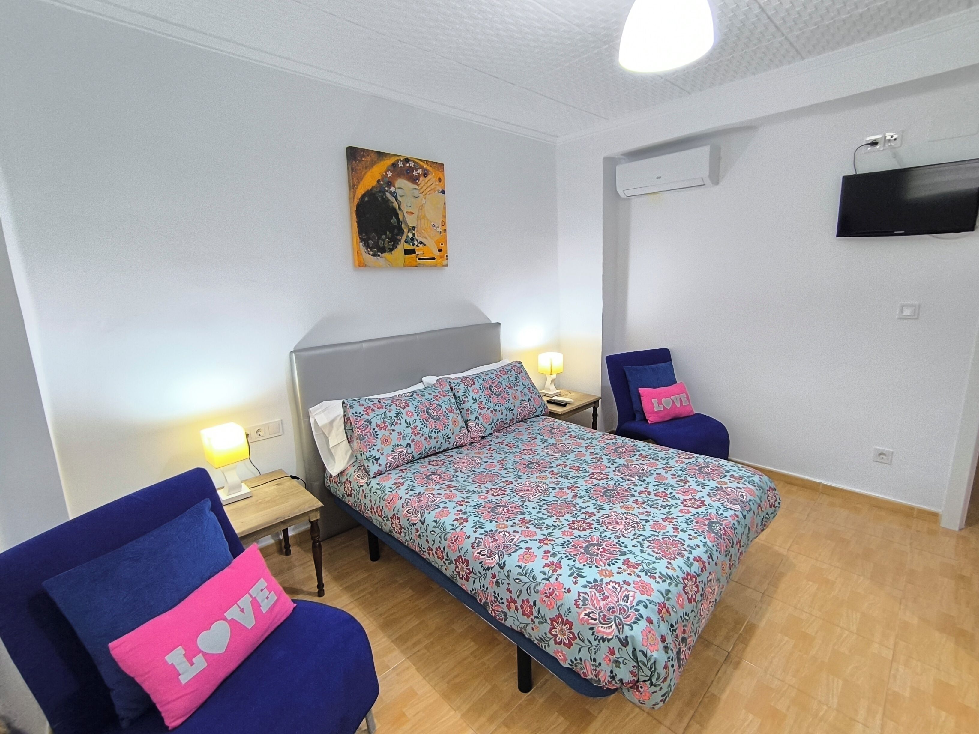 A bedroom with a bed, two chairs, a nightstand, a lamp, a painting, and a TV.