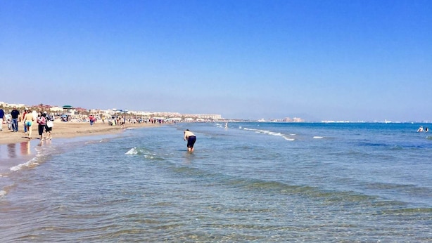 A beach with clear shallow waters, a person wading in the water, and a distant shoreline with buildings. - Apartment 2 rooms for rent for more than 10 days with free parking. (València)