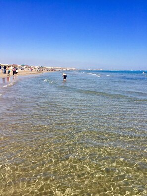 Una platja amb aigües poc profundes i cristal·lines, una persona caminant a l'aigua i una riba llunyana amb edificis.