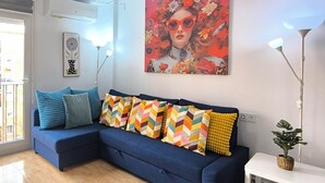 A modern living room with a blue sectional sofa, colorful pillows, a large floral painting, and a potted plant on a white console table. - Apartment 2 rooms for rent for more than 10 days with free parking. (València)