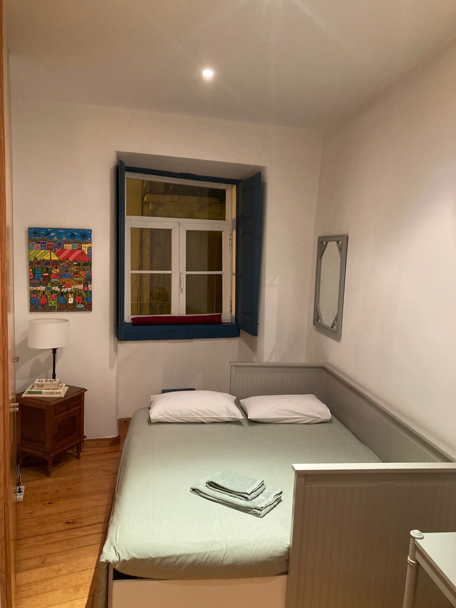 Ein Schlafzimmer mit einem Bett, einem Fenster mit blauen Fensterläden, einem Nachttisch mit Lampe und einem farbenfrohen Bild an der Wand.