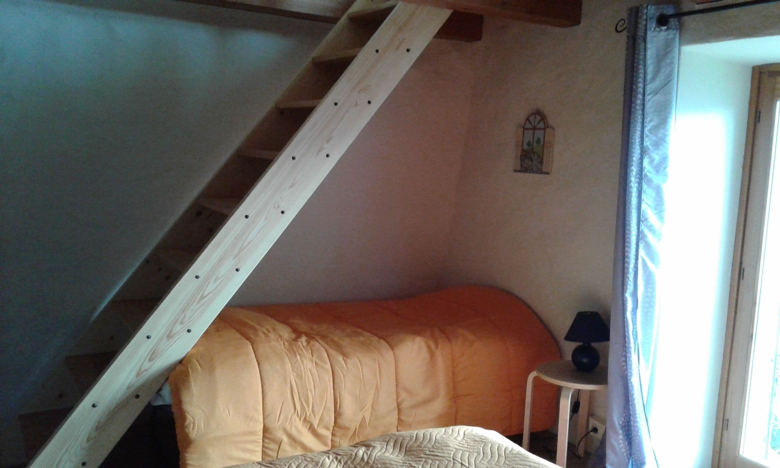 Ein Schlafzimmer mit schrägverdeckter Decke, einem Einzelbett, einem kleinen Tisch mit Lampe und einem Fenster mit Vorhängen.