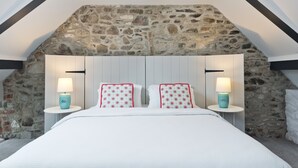 Un dormitorio con pared de piedra, techo inclinado y cama con ropa de cama blanca y almohadas estampadas. Hay dos mesitas de noche con lámparas.