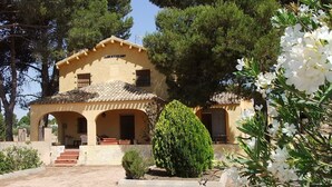 Una villa llamada 'Villamaría' con techo de tejas, entrada en arco y un jardín con un arbusto cuidadosamente recortado y flores blancas.