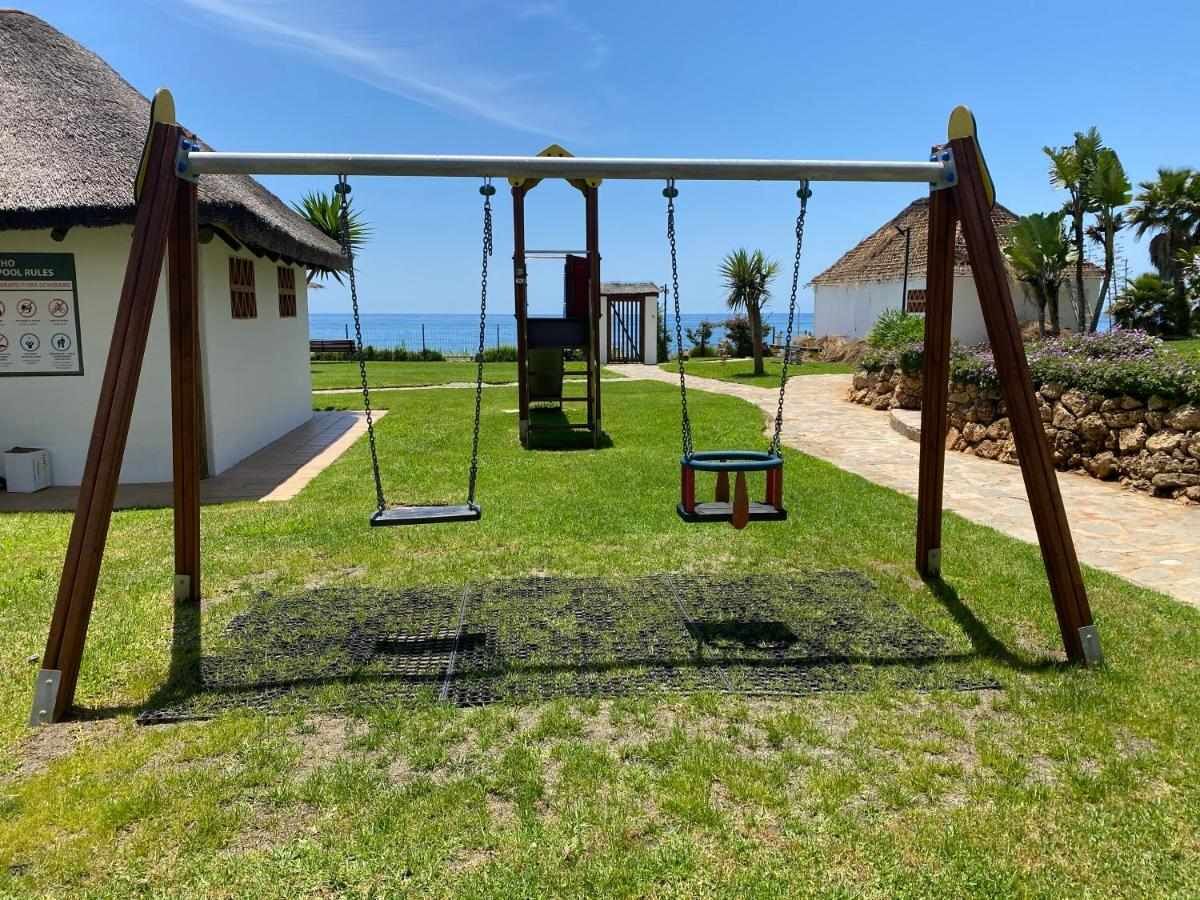 A playground with swings, a thatched-roof structure, and a view of the ocean.