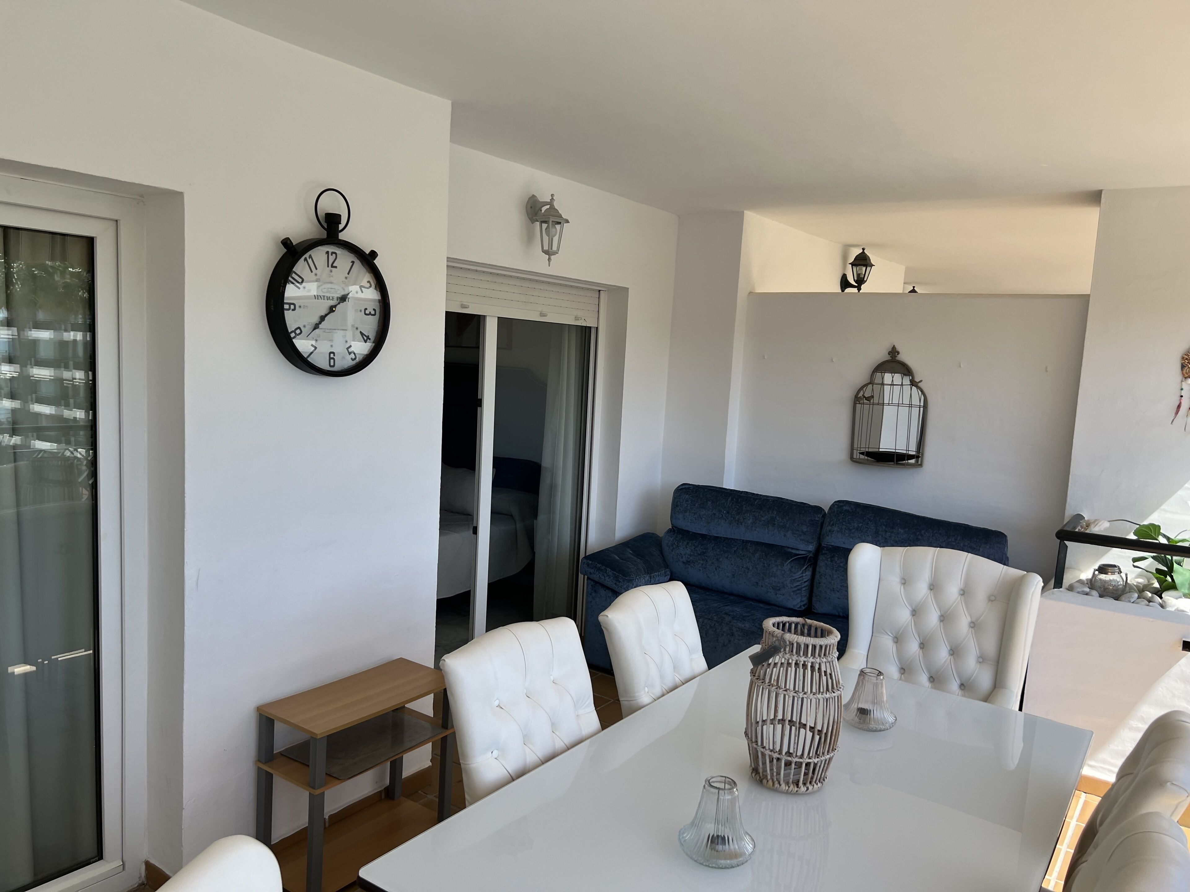 A modern living room with a white sofa, a dining table with chairs, a clock, and a sliding glass door leading to another room.