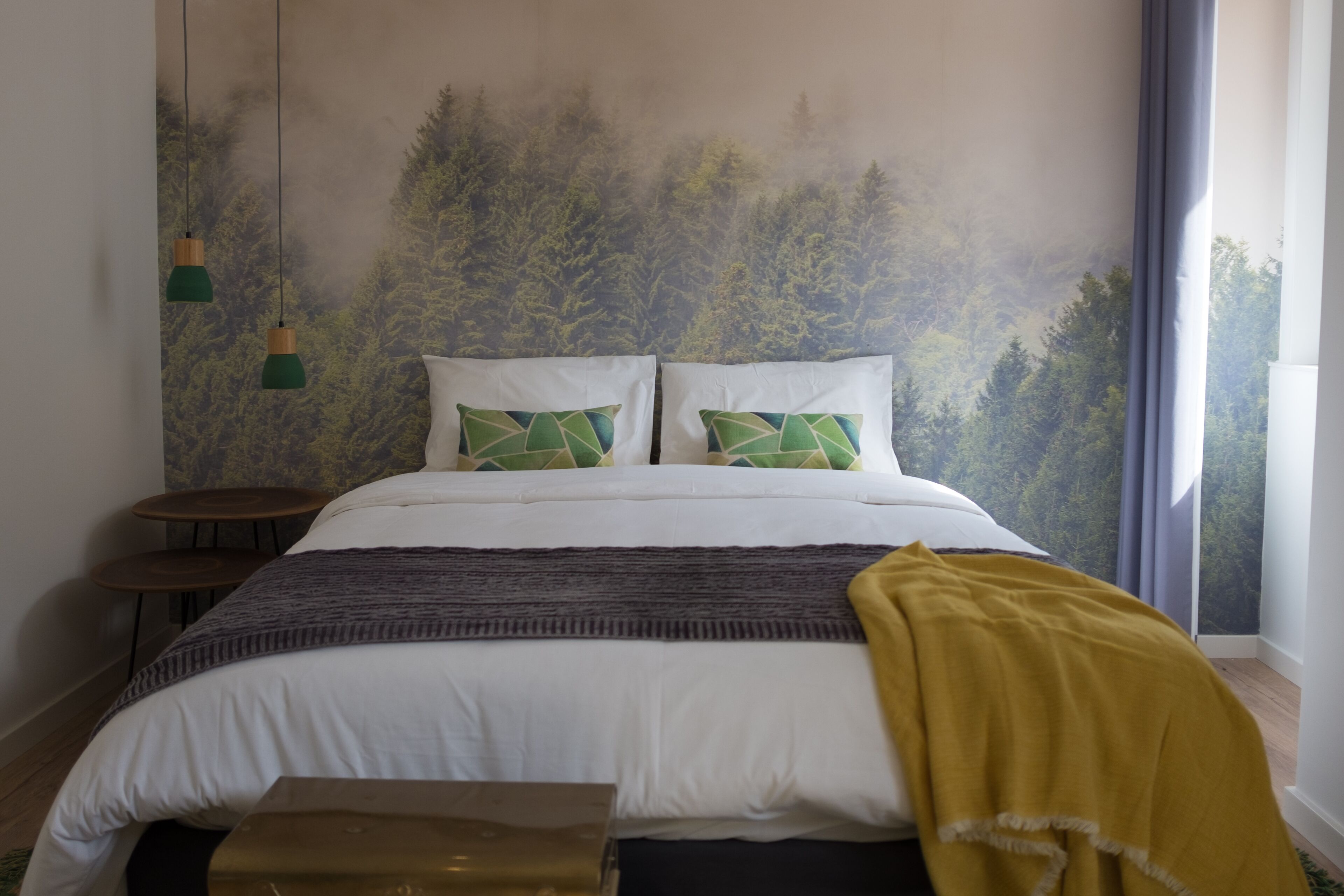 Un dormitorio con cama, dos almohadas, una colcha, una mesita y un mural en la pared.