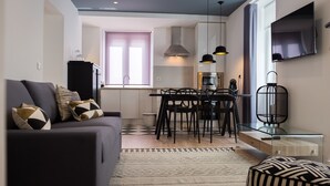 A modern living room with a sofa, dining table, chairs, and a TV mounted on the wall. - Ocean Center Apartments - Ocean Green (Cascais)