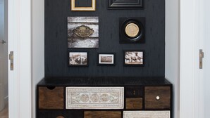 A dark wooden wall with a round mirror, framed pictures, and a decorative welcome sign. Below, there is a wooden console with drawers and a decorative tray. - Ocean Center Apartments - Ocean Green (Cascais)