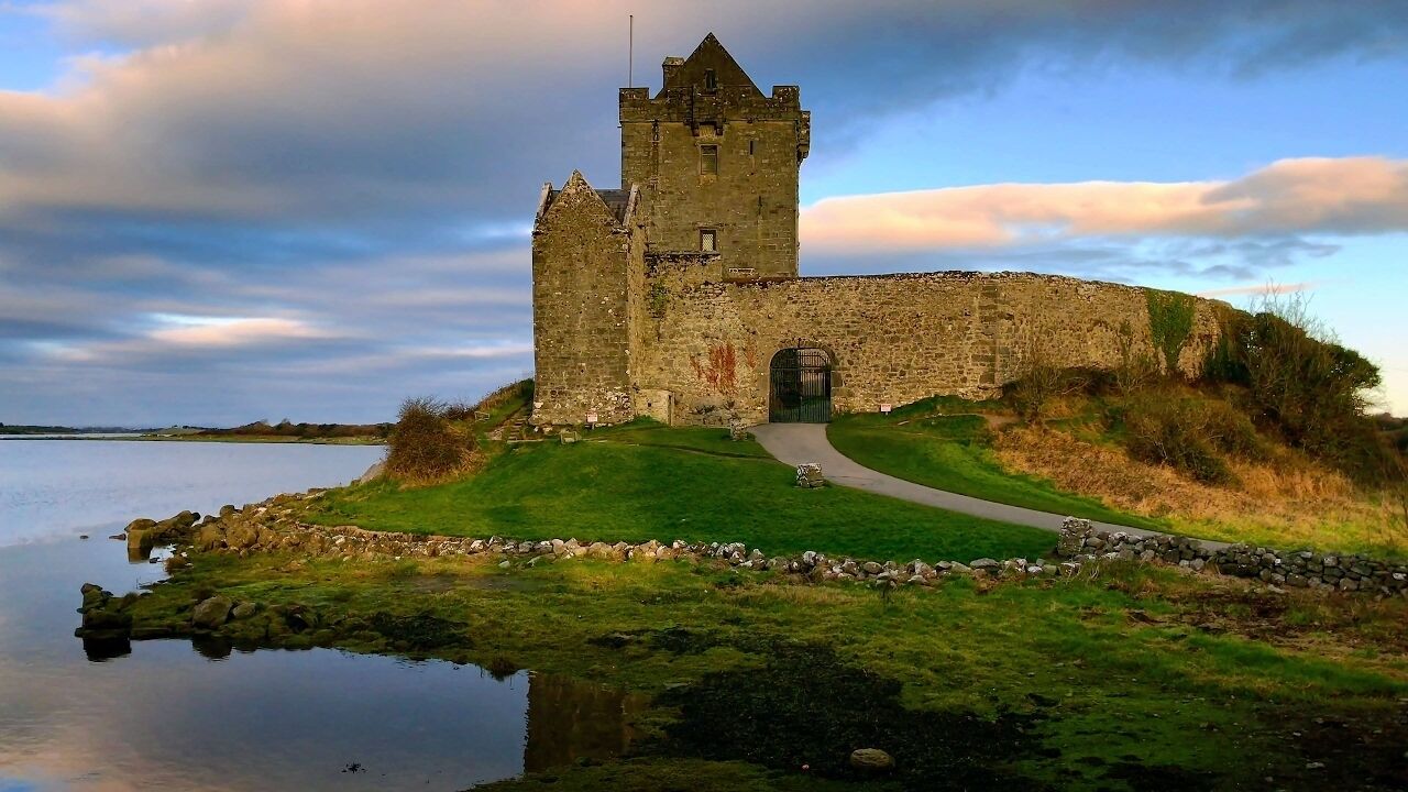 A historic stone castle with a central tower, a paved pathway leading to the entrance, and a body of water reflecting the structure.