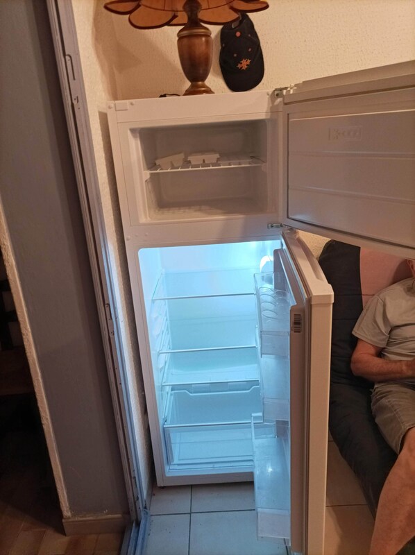 Un refrigerador con la puerta abierta, mostrando estantes vacíos y una persona sentada a su lado.
