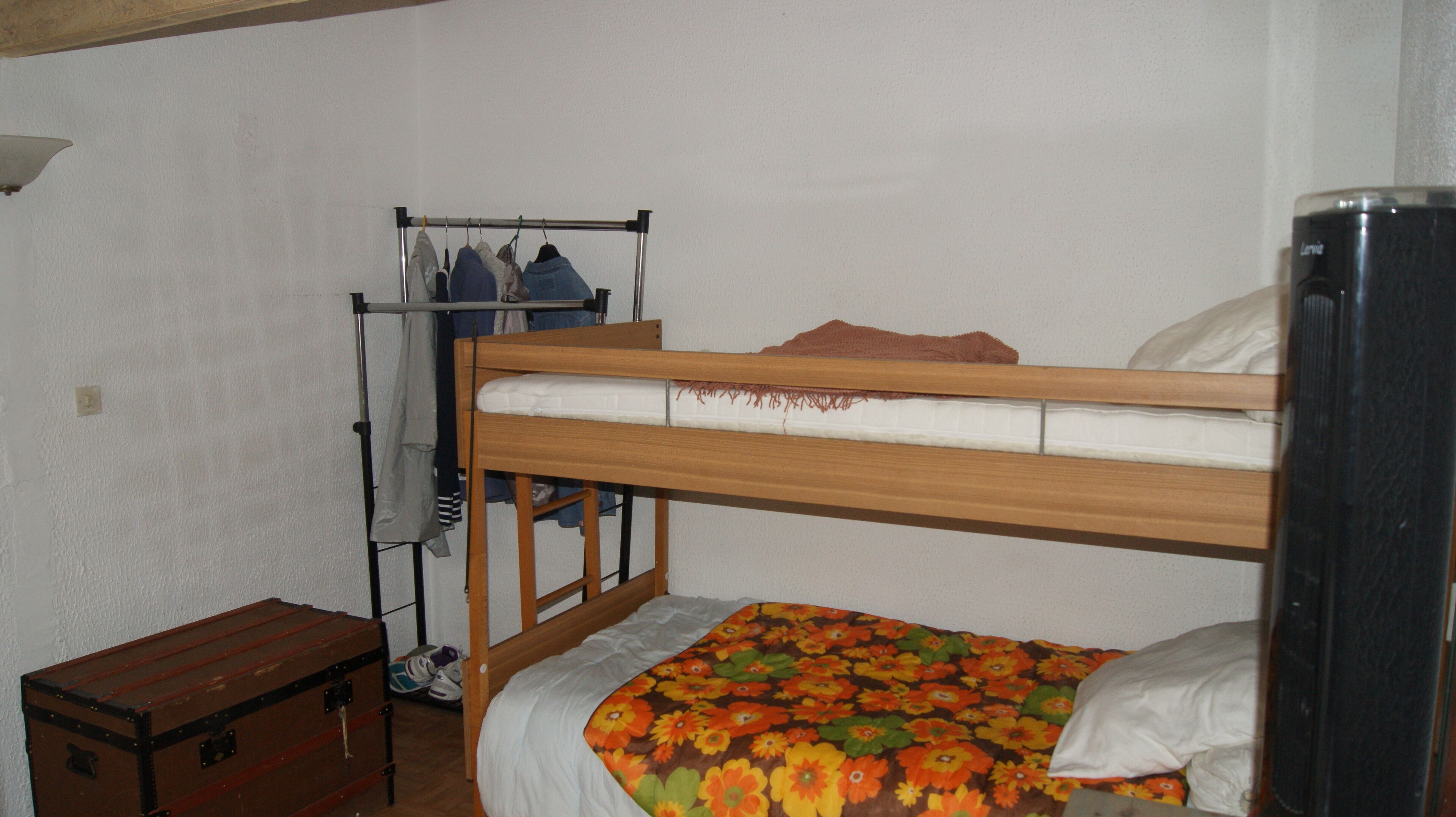 Une chambre avec un lit simple, un cadre en bois, une couvre-lit à motifs floraux, une petite commode en bois, un portant à vêtements et un réfrigérateur noir.