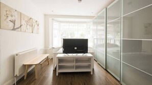 Une pièce moderne avec un radiateur blanc, un banc en bois, un téléviseur posé sur un meuble blanc et une grande fenêtre équipée de stores.