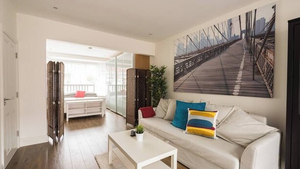 Un salon moderne avec un canapé blanc, des coussins colorés, une table basse blanche et un grand tableau représentant un paysage urbain accroché au mur.