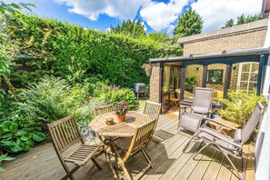 Une terrasse en bois avec une table et des chaises, entourée d’une végétation luxuriante et d’un bâtiment en briques.