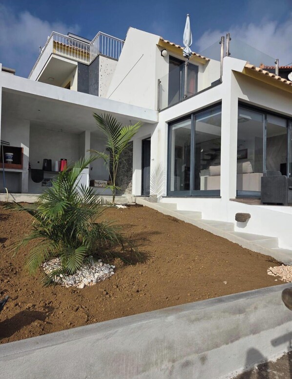 A modern house with a flat roof, large glass windows, and a small garden with palm trees. - House Crystal Calheta (Calheta (Madeira))