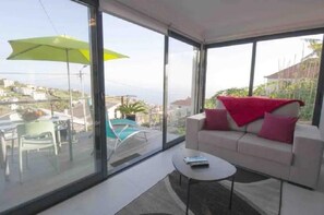 Un salon moderne avec un canapé, une table basse et une grande fenêtre donnant sur la mer et les bâtiments environnants.