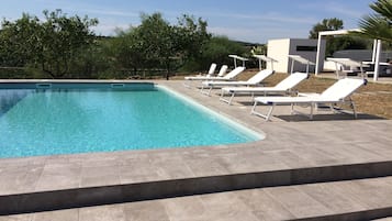 Una piscina con acqua cristallina, circondata da un solarium piastrellato e poltrone da relax bianche.