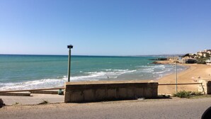 Une route côtière bordée d’une plage et de l’océan sur la droite, une barrière en béton au premier plan et un ciel bleu dégagé.