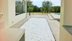 Une terrasse extérieure moderne, avec un chemin en petits galets blancs, un petit arbre et une piscine en arrière-plan.