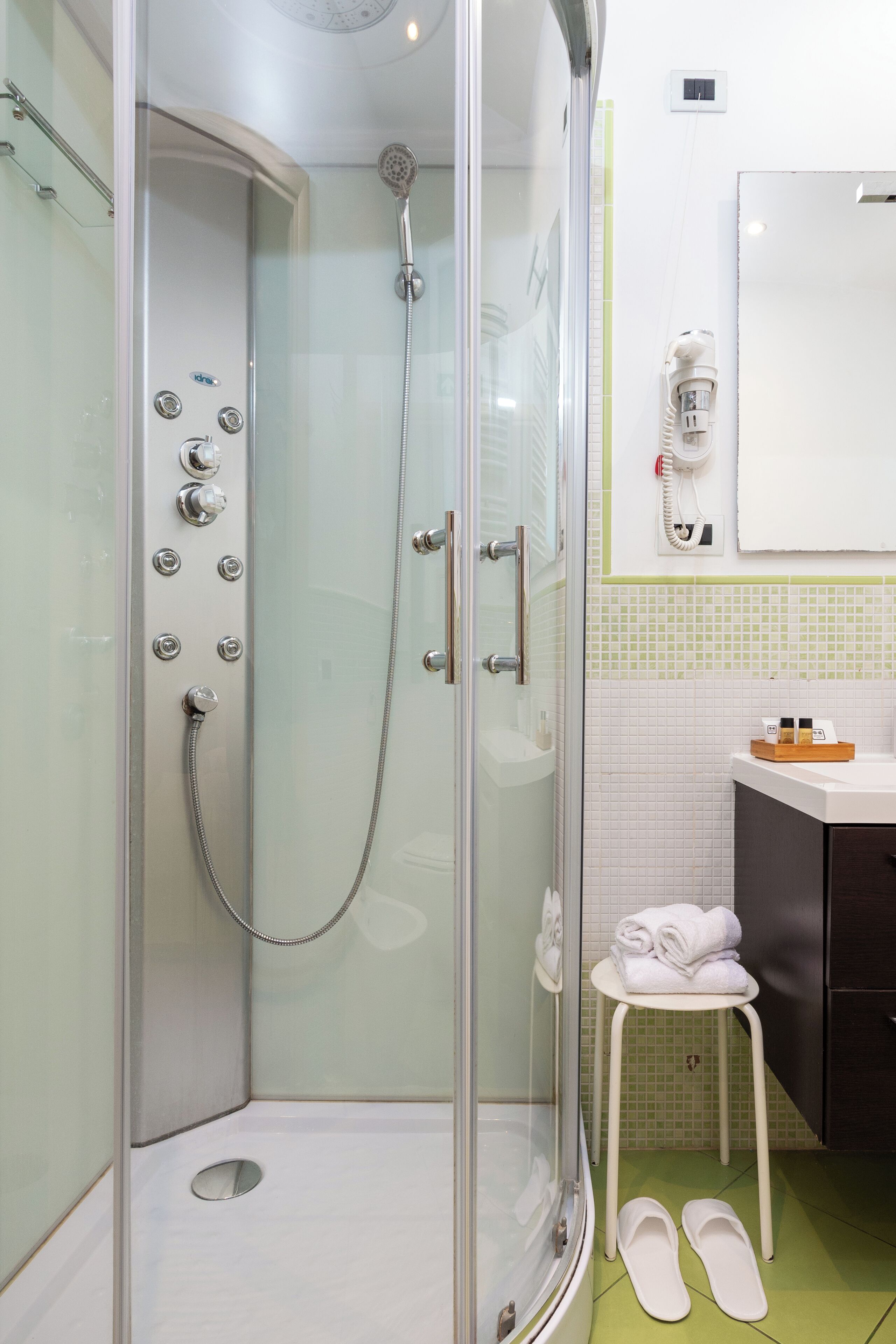 A glass-enclosed shower with multiple controls, a white toilet, and a small stool with a towel.