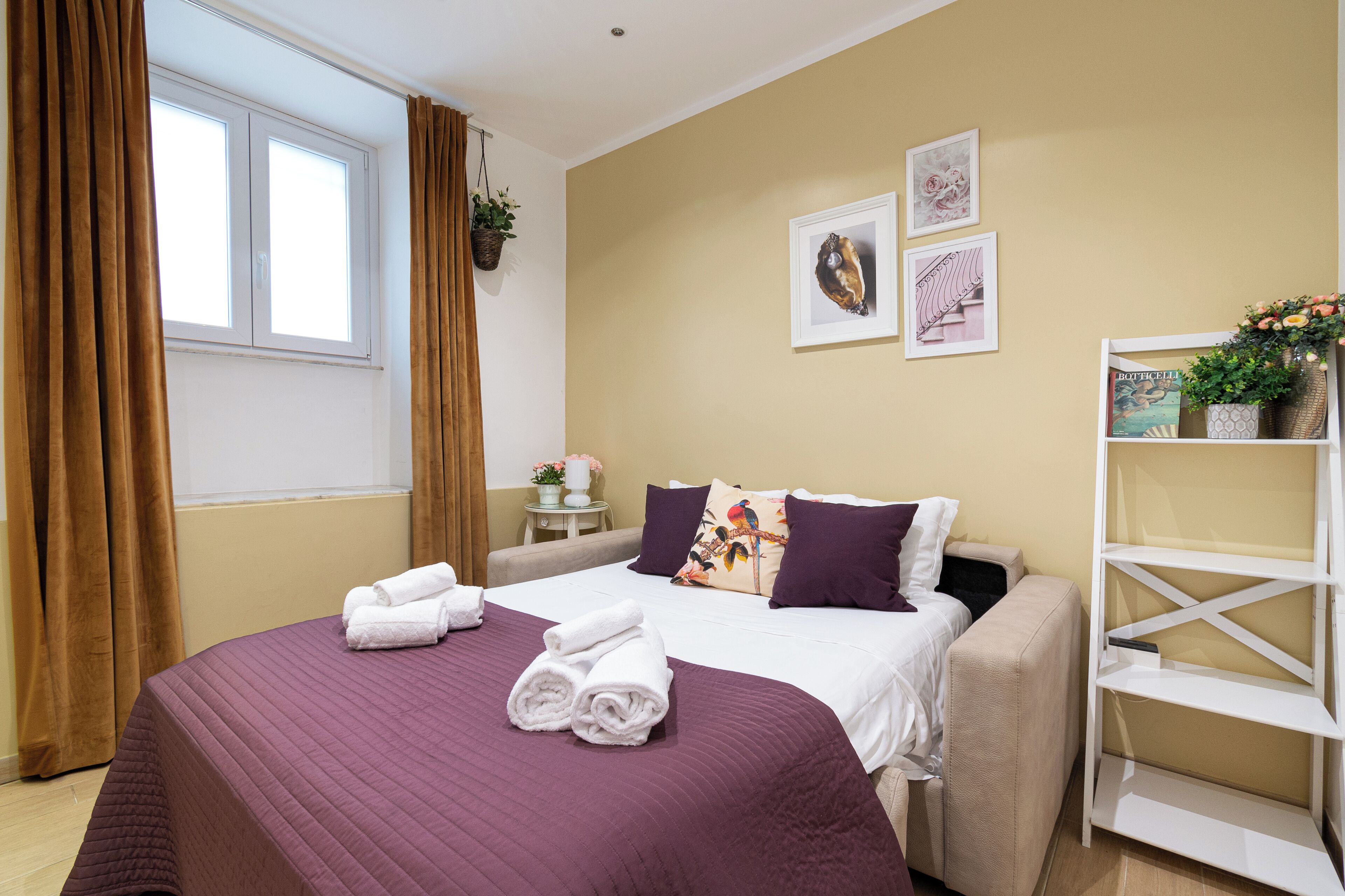 A neatly made bed with a purple bedspread and two rolled towels, a white bedside table with a plant, and a wall with two framed pictures.