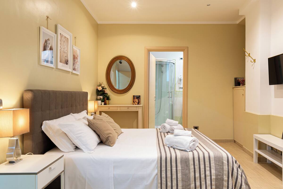 A hotel room with a bed, bedside table, lamp, and a shower area.