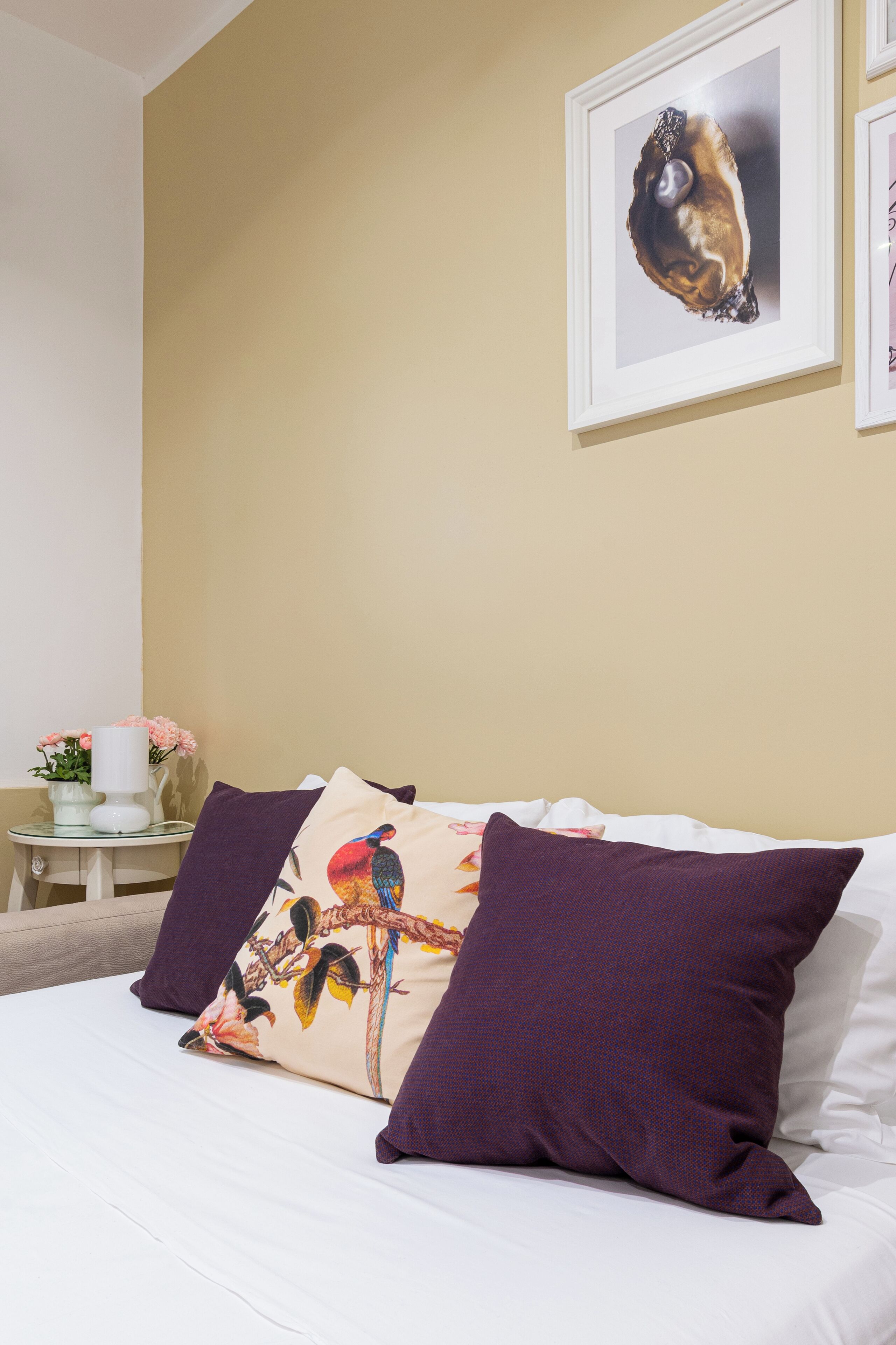 A neatly made bed with white linens and two purple pillows, a framed artwork above the bed, and a small table with a vase of flowers to the left.