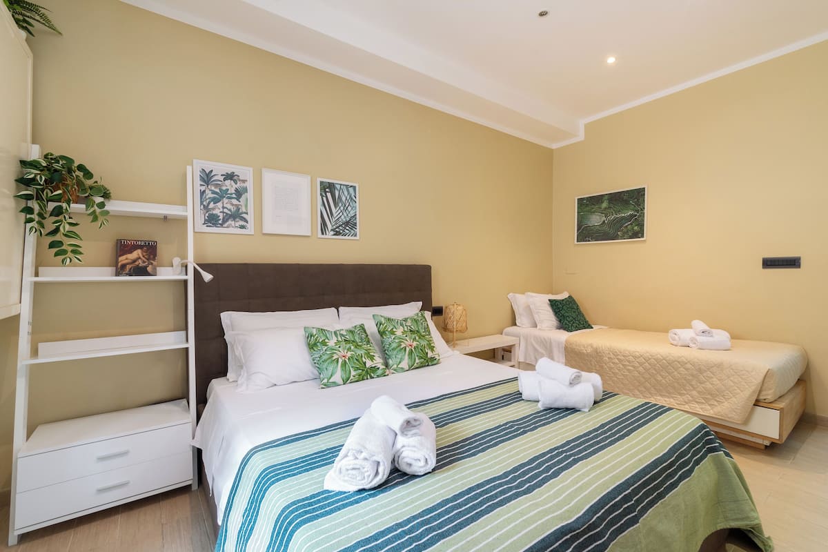 A hotel room with a bed, a nightstand, a shelf with plants, and artwork on the walls.