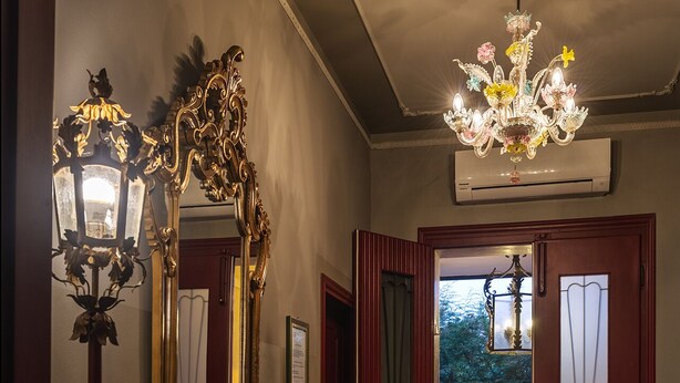 A hallway with a wooden dresser, a large ornate mirror, and a chandelier. There is a door leading to an outdoor area with a chair and a lantern.
