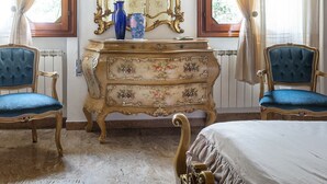 A bedroom with a large ornate mirror, two blue upholstered chairs, a wooden dresser, and a bed with a white bedspread.