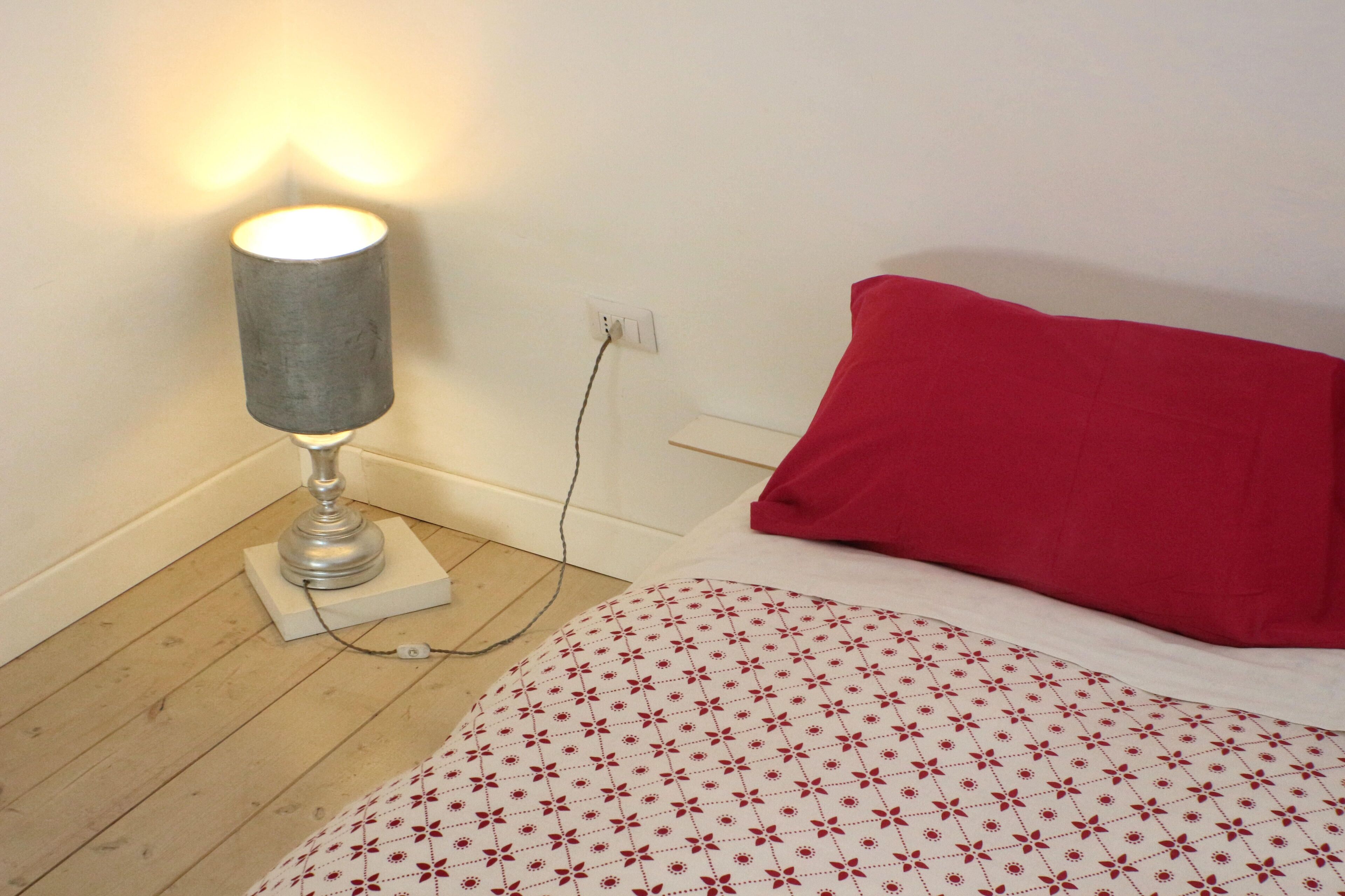 A room with a bed, a red pillow, a patterned bedspread, a lamp, and a power outlet on the wall.